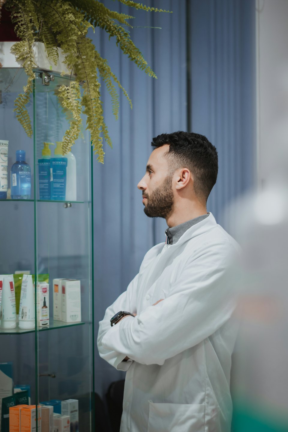 Pharmacist in white coat examines products in glass cabinet.